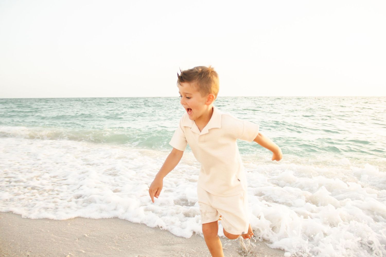 Joyful person in white linen walking along serene beach with waves breaking at sunset.