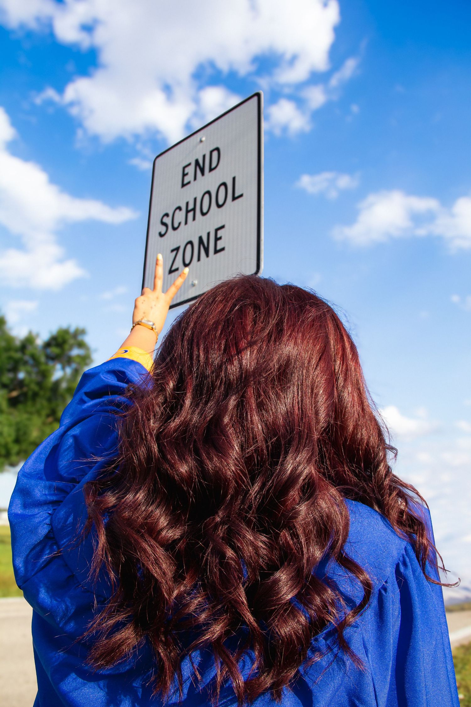 A graduate in blue gown touches an End School Zone sign, celebrating completion of school under a blue sky.