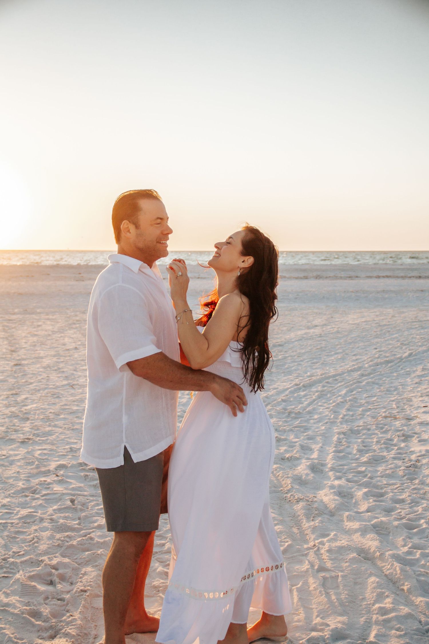 Couple embracing and smiling at sunset on a sandy beach, woman in white dress, man in white shirt and gray shorts.