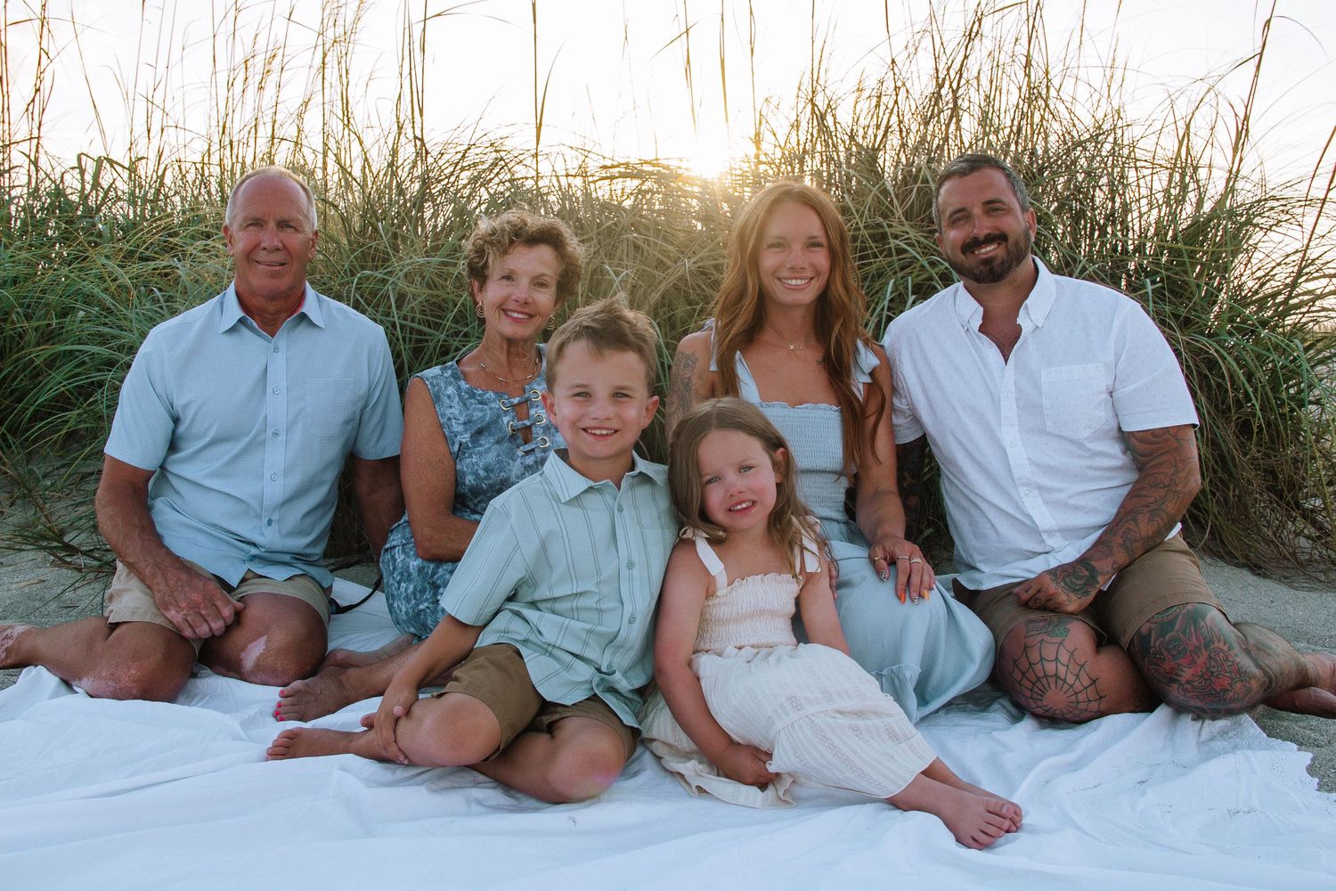 Family of six poses on a white blanket at a sandy beach with golden sunset light and tall grass behind them.