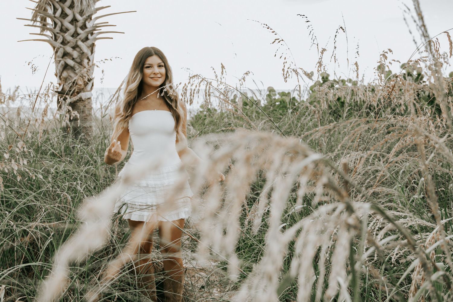 Young woman in white dress and brown boots standing among tall golden grasses near a palm tree outdoors.