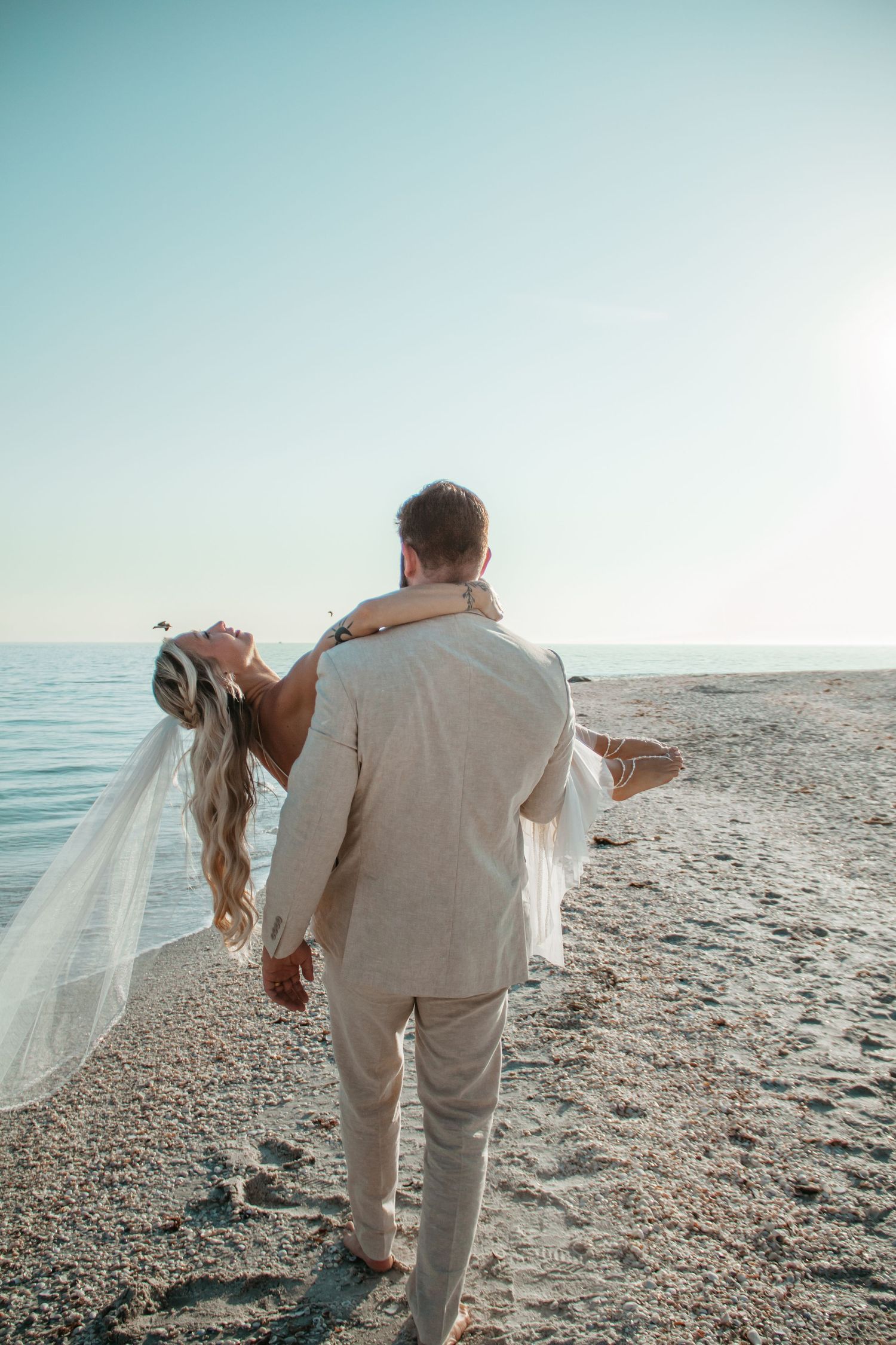 Groom carrying bride in white wedding dress on a sandy beach with calm ocean and clear blue sky.