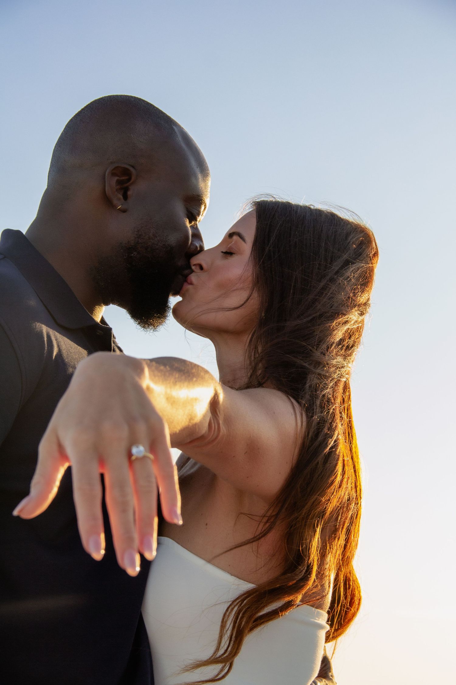 Interracial couple sharing a romantic kiss while showing off an engagement ring against a blue sky backdrop.