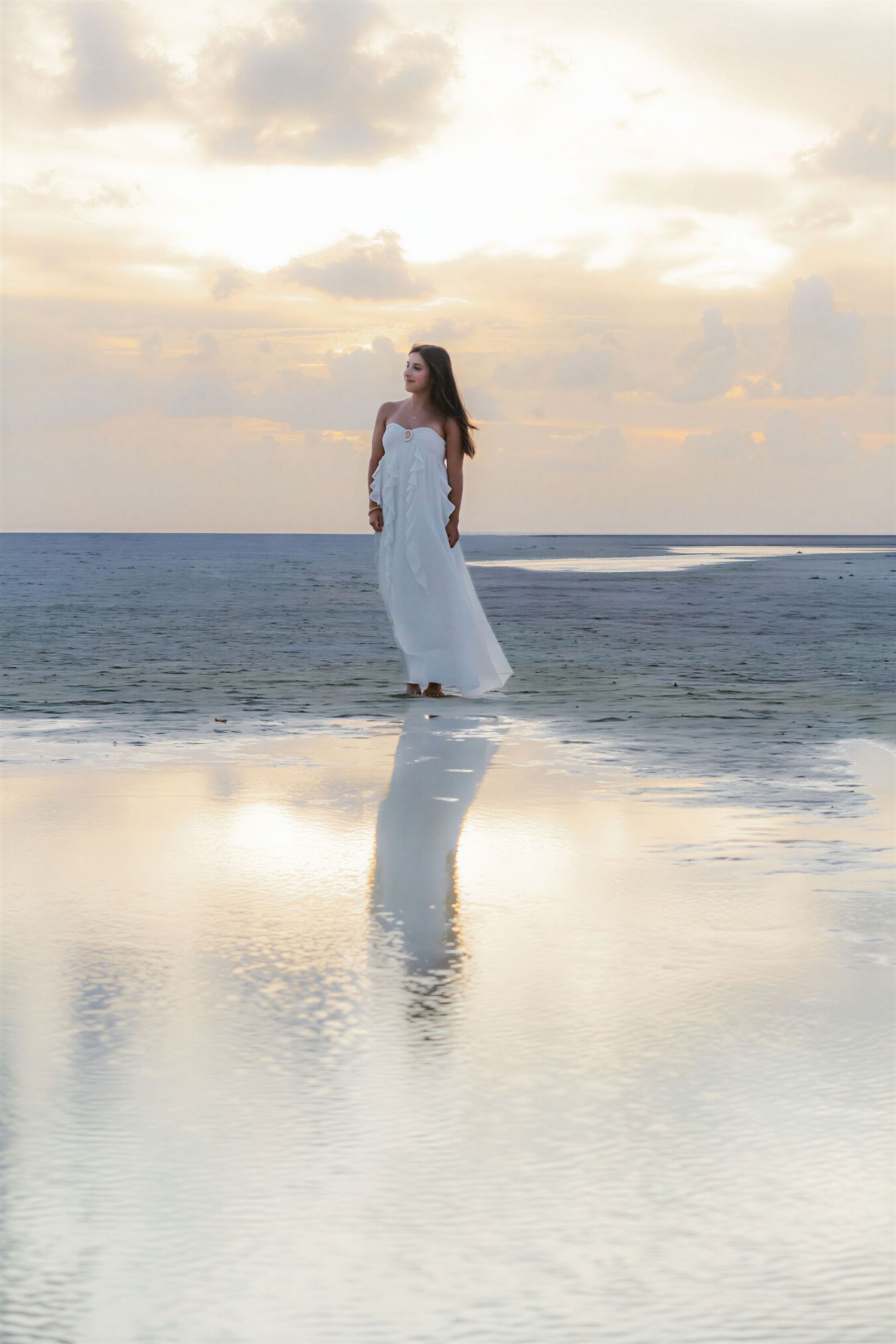 Woman in white dress standing on a reflective beach at sunset with ocean horizon backdrop.