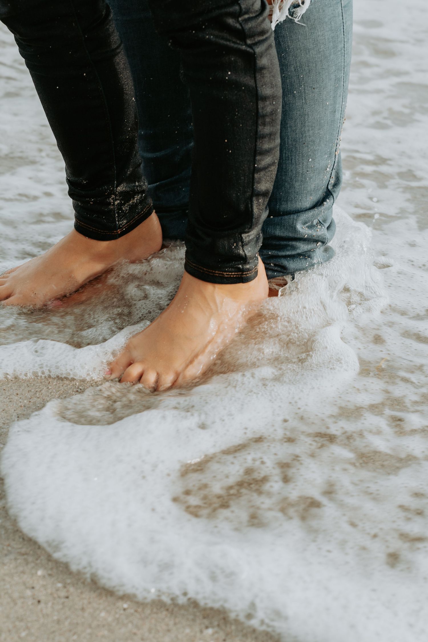 Two people standing barefoot in ocean waves on a sandy beach, wearing rolled-up jeans as foam washes over their feet.