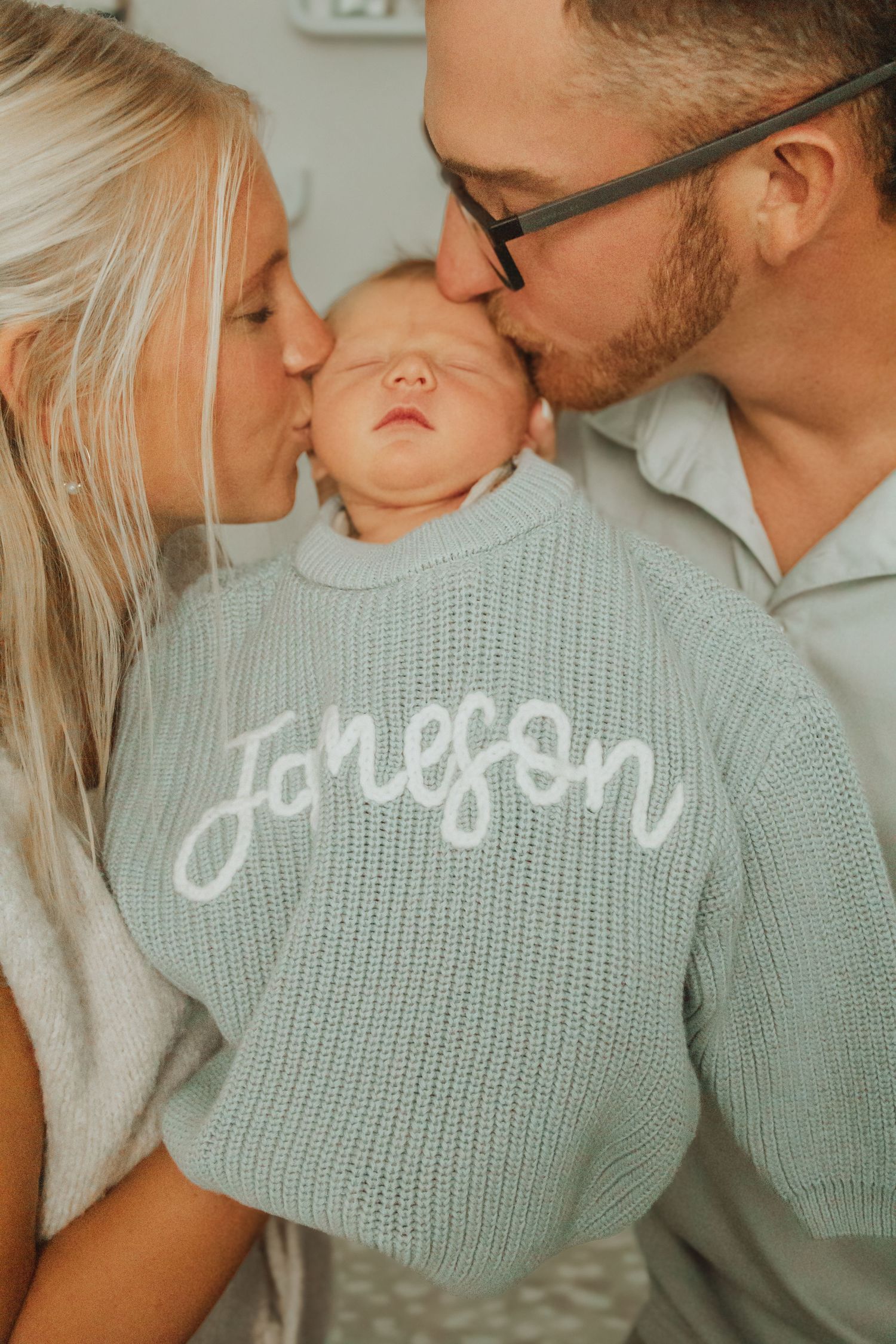 Parents kissing newborn baby wearing a mint green knit sweater with the name Joneson embroidered on the back.