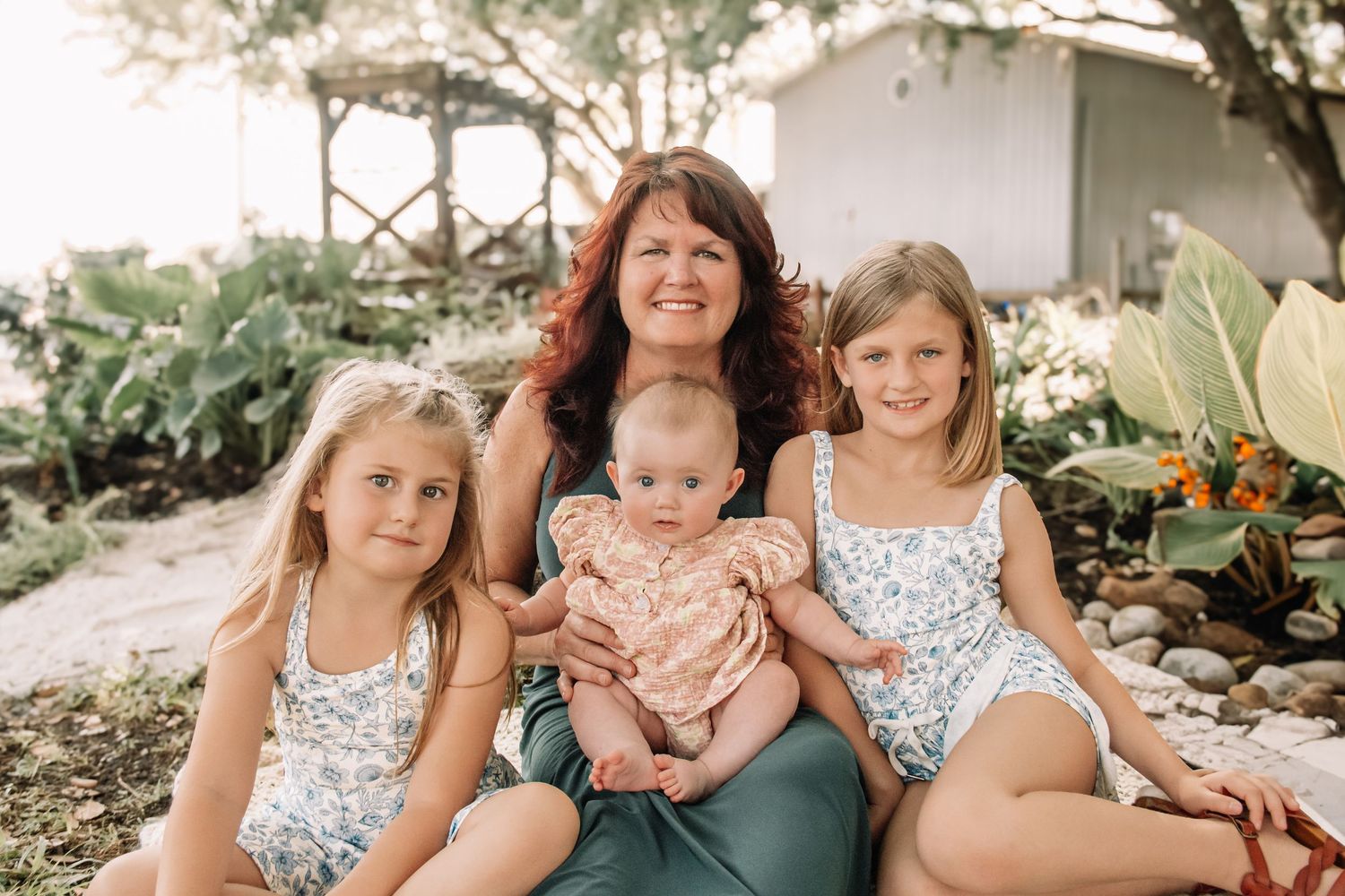 A smiling grandmother with curly red hair poses outdoors with three young granddaughters, including a baby, in floral outfits.
