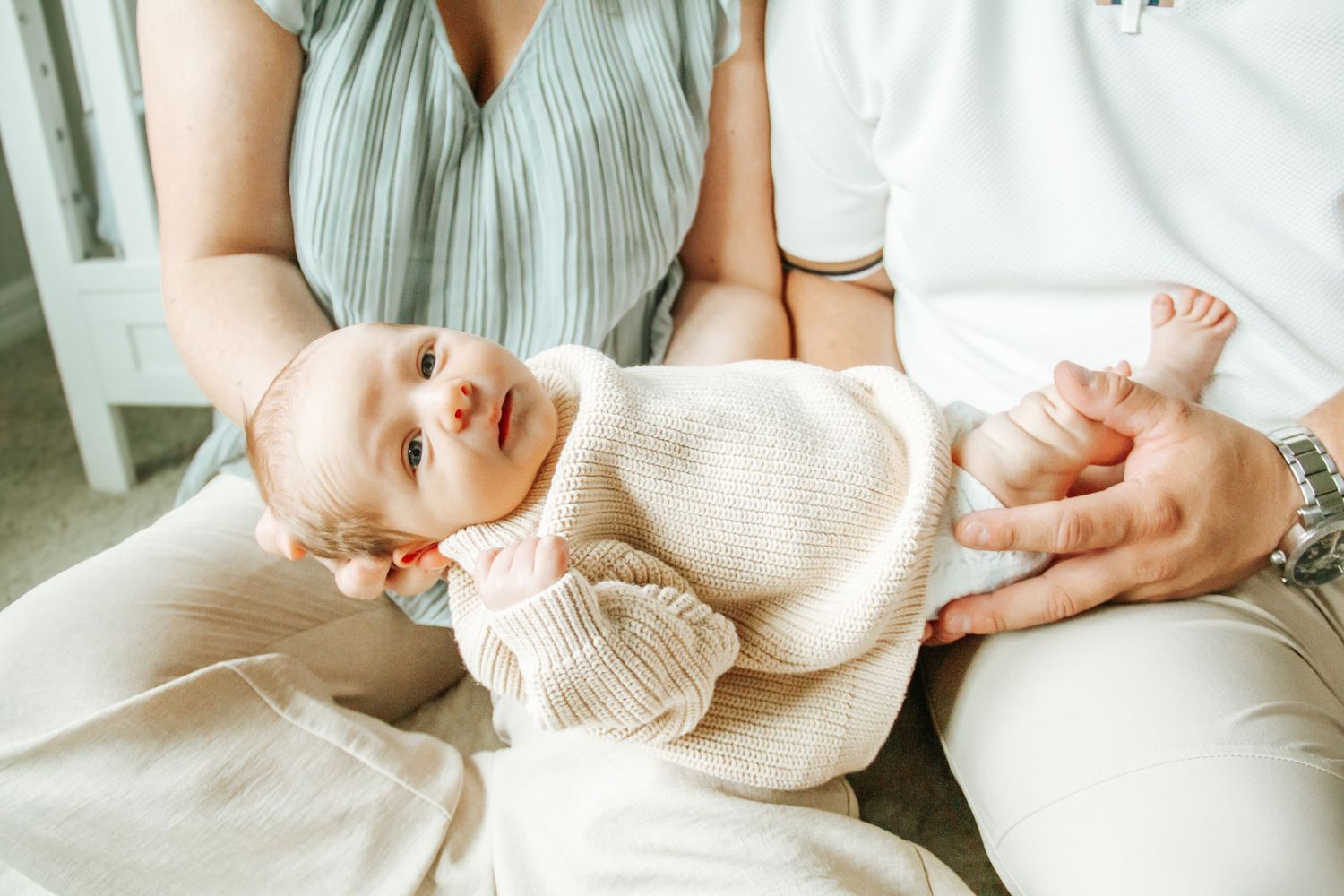A newborn baby in a cream knit sweater rests in the arms of two parents wearing white and mint outfits.