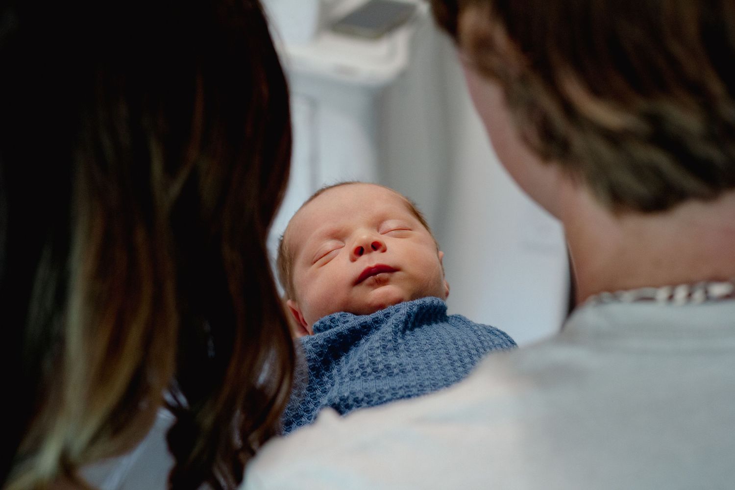 Two adults lovingly hold a sleeping newborn baby wrapped in a blue knit blanket in a hospital room.