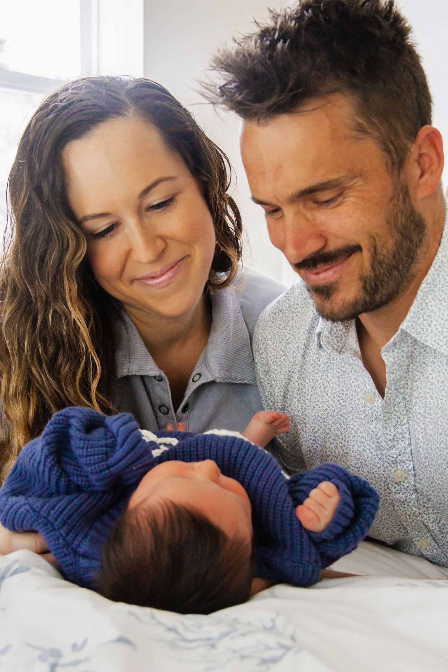Smiling couple gazing lovingly at their newborn baby dressed in a blue knit outfit lying between them.