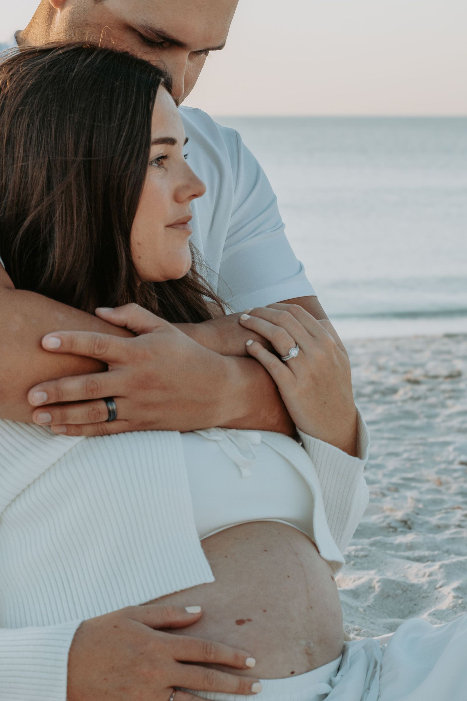 Couple embracing on a sandy beach at sunset, woman wearing engagement ring, man hugging her from behind near ocean waves.