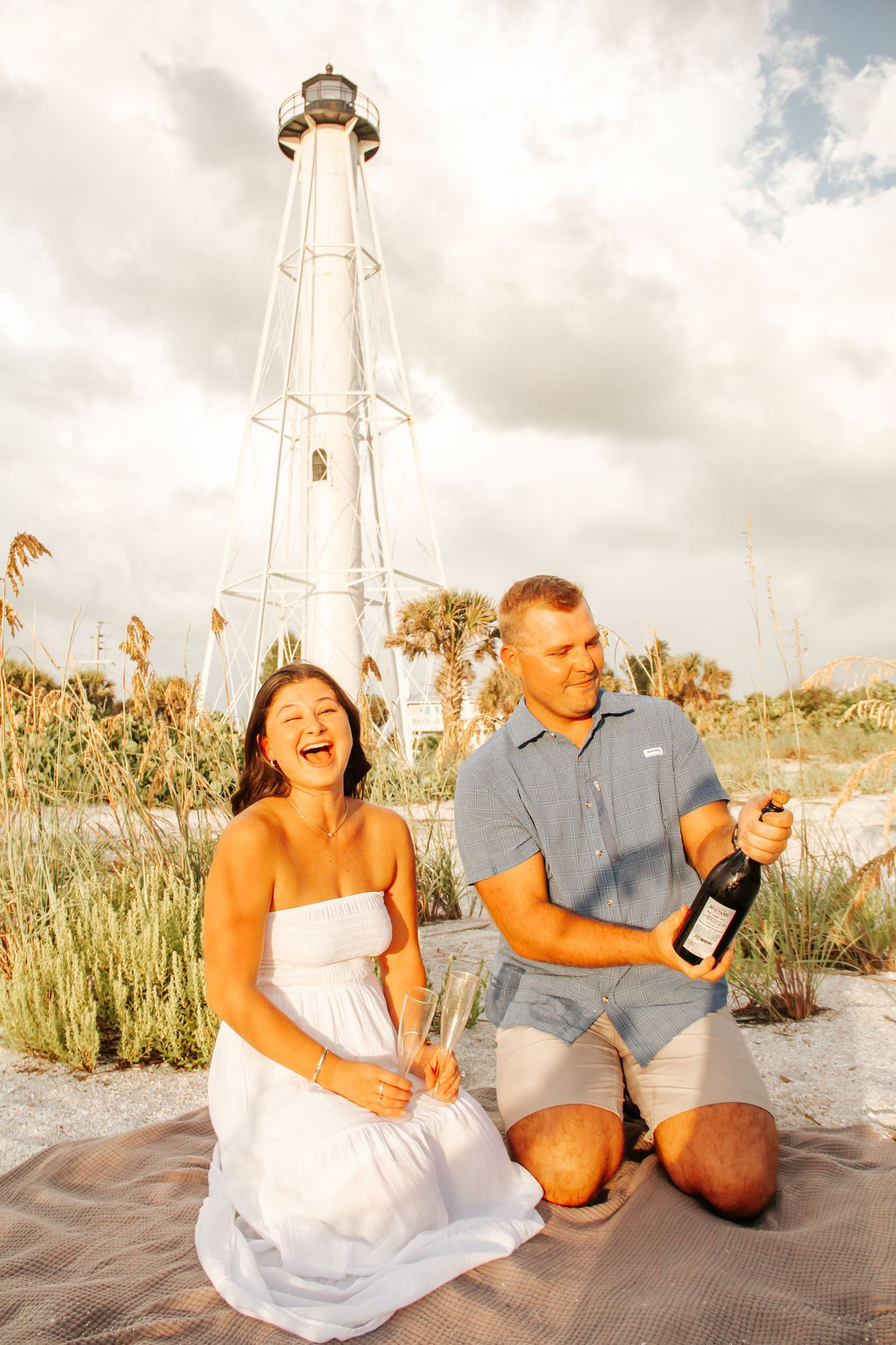 Couple celebrating with champagne on beach near white lighthouse, woman in white dress laughing joyfully.
