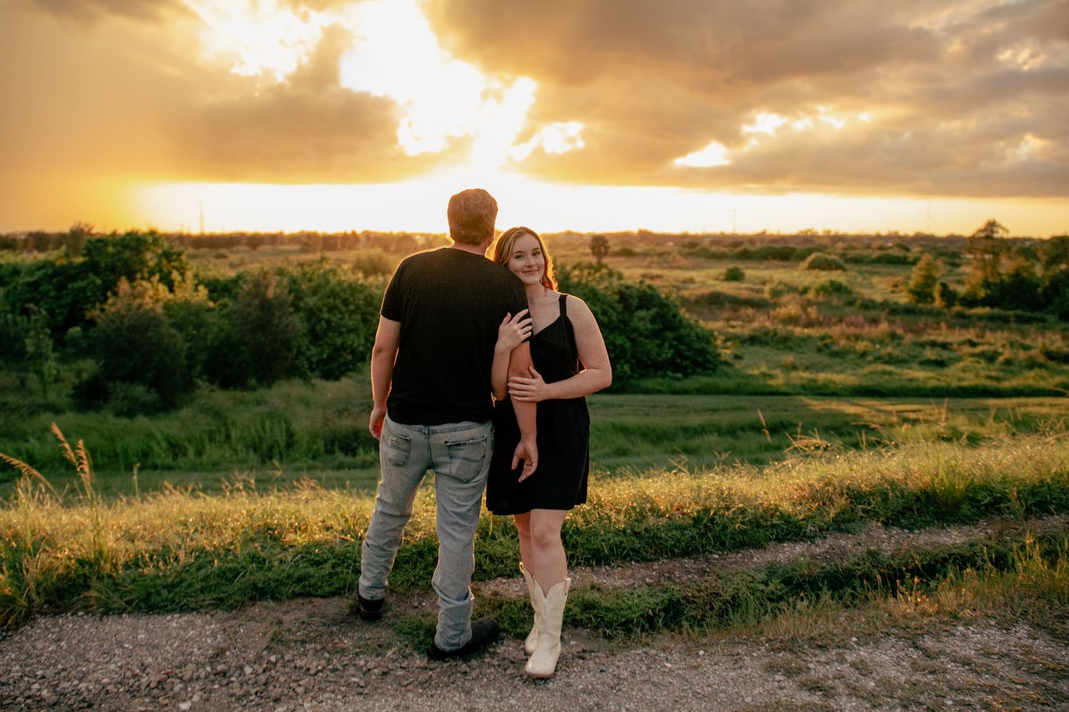Couple embracing at sunset overlooking a scenic green landscape, woman in black dress smiling warmly at golden hour.