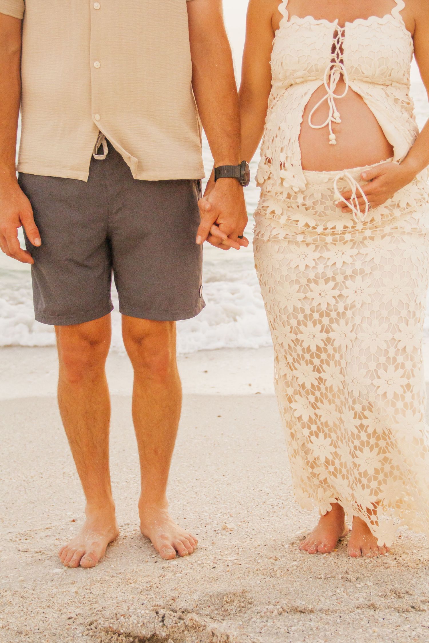 Couple holding hands on sandy beach, woman in lace dress showing baby bump, man in gray shorts and beige shirt.