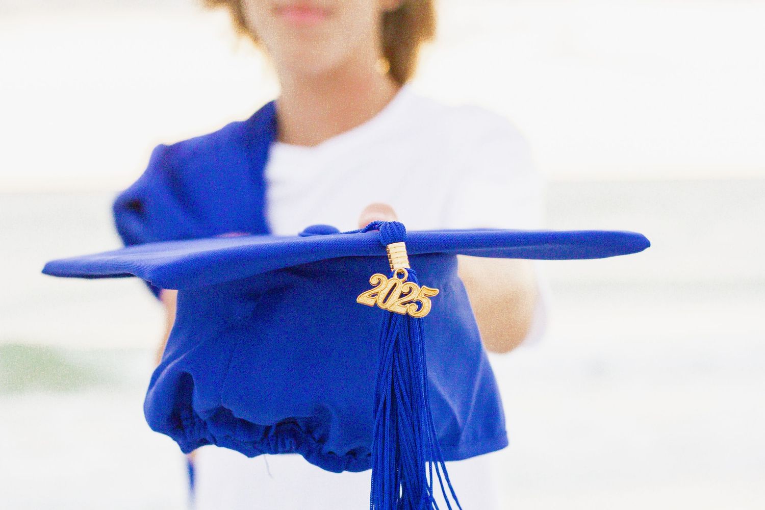 A blue graduation cap with a gold 2025 tassel charm held out by a graduate in a white shirt.