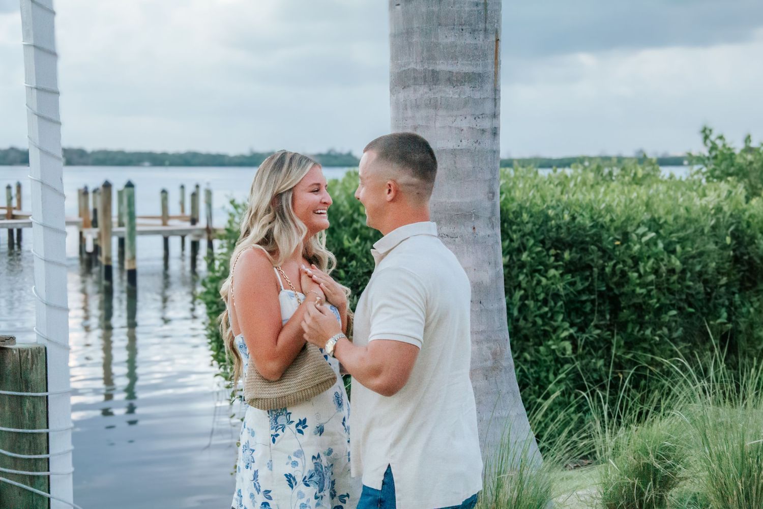 Couple sharing a romantic moment by a waterfront dock surrounded by lush greenery on a cloudy day.