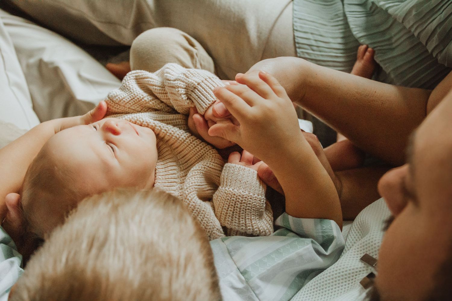 Newborn baby in knit outfit lying on white bedding while older sibling gently holds tiny hand.