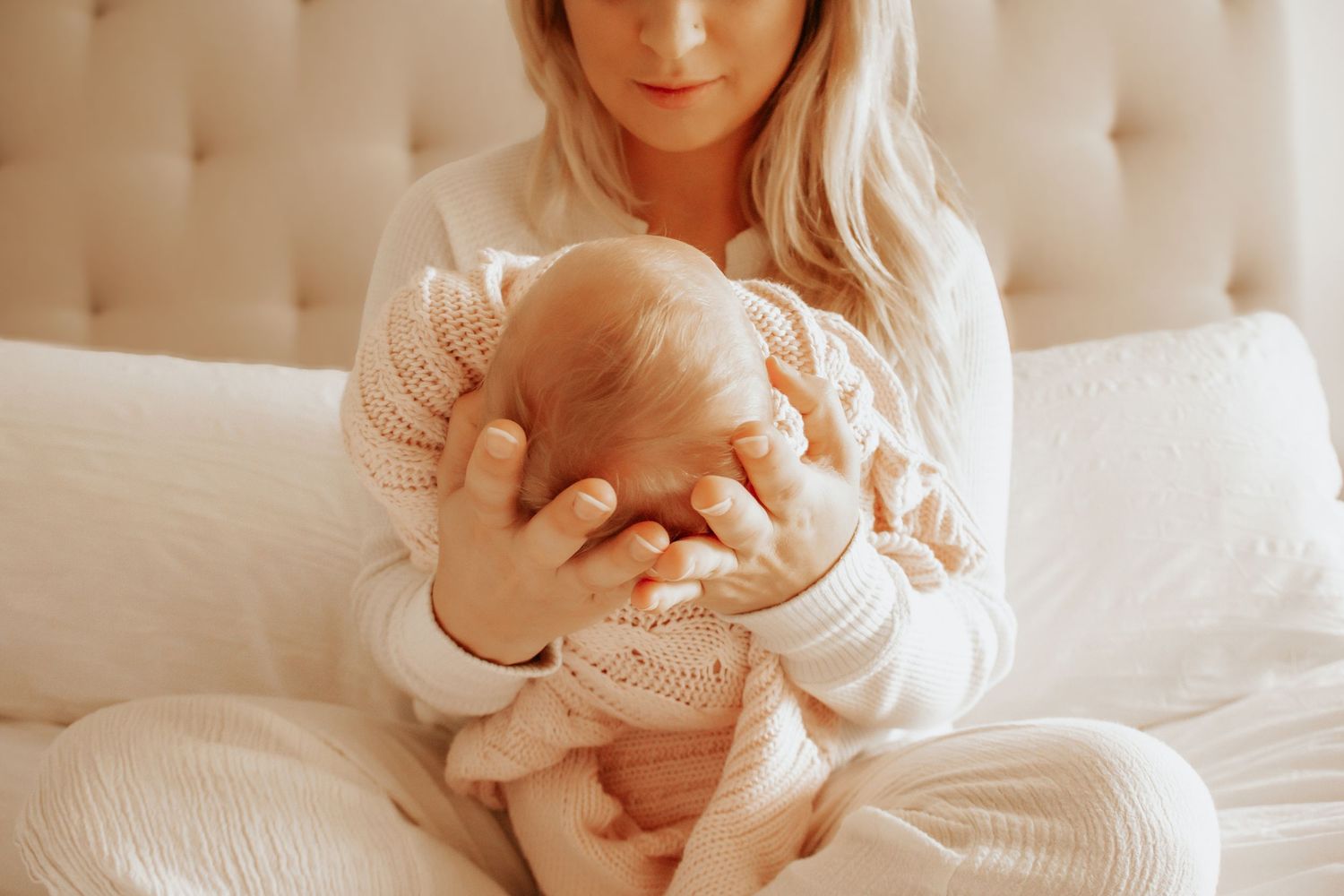 A blonde mother in white pajamas tenderly holds her newborn baby while sitting on a bed.