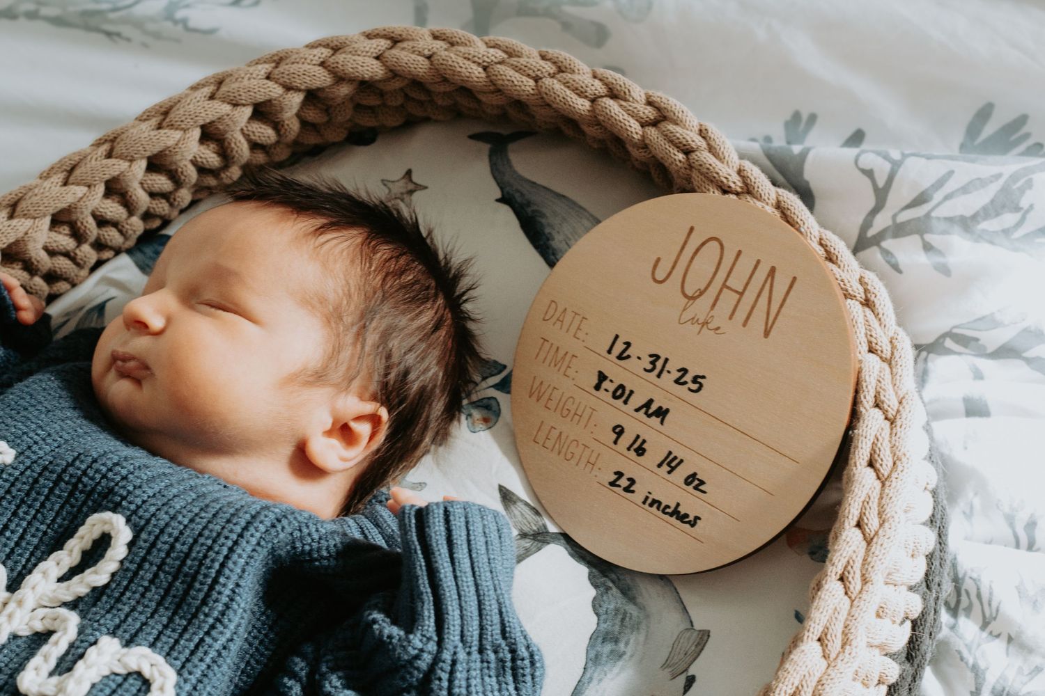 Newborn baby John sleeping in a knit basket beside a wooden birth announcement sign showing stats.