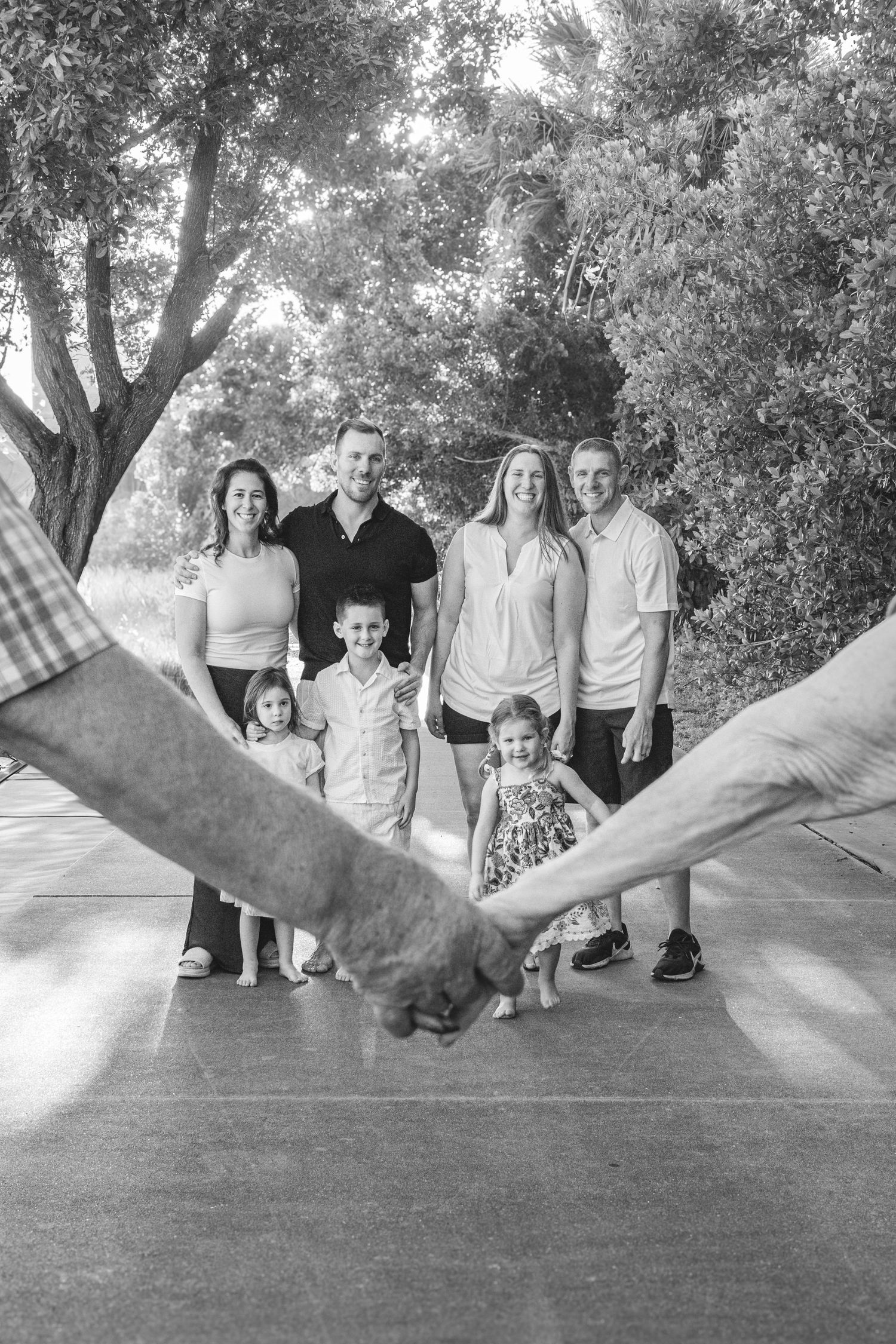 Black and white family portrait of two couples with three young children holding hands outdoors near trees.