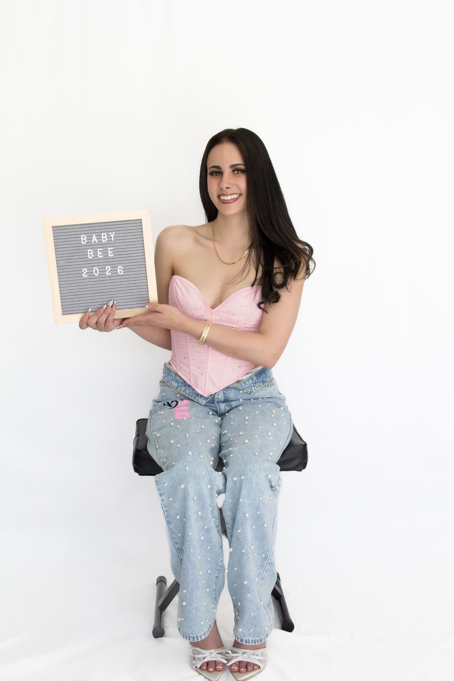 Smiling woman in pink corset and jeans sits holding a letter board sign reading Baby Bee 2026.
