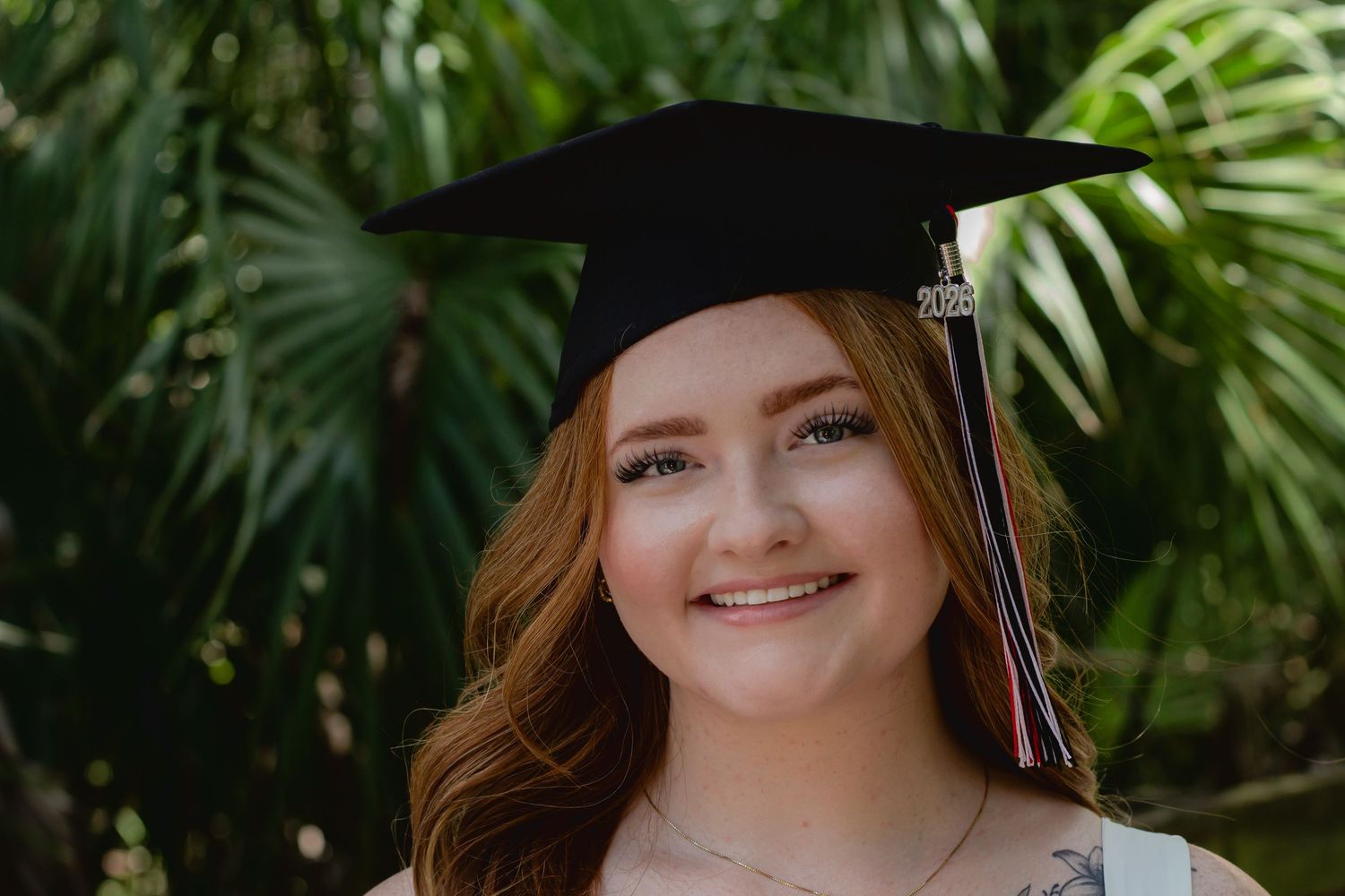 Smiling young woman wearing a black graduation cap with tassel, posing outdoors with tropical greenery background.