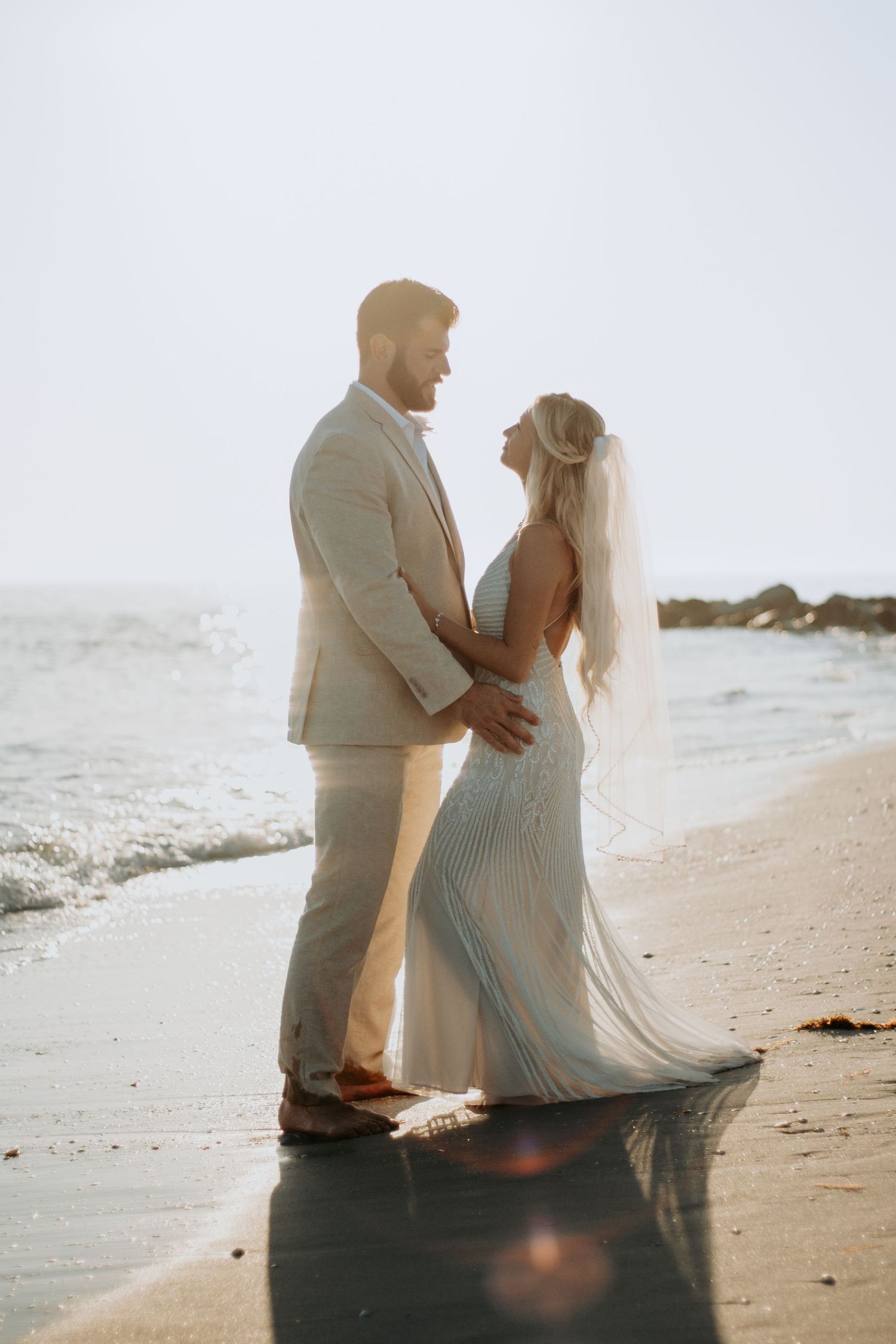 Bride and groom share a romantic beach moment at sunset, with waves and golden light surrounding them.