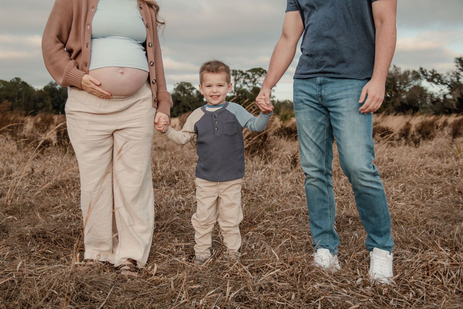 Pregnant woman and man holding hands with young boy in a field during a fall maternity photo session outdoors.