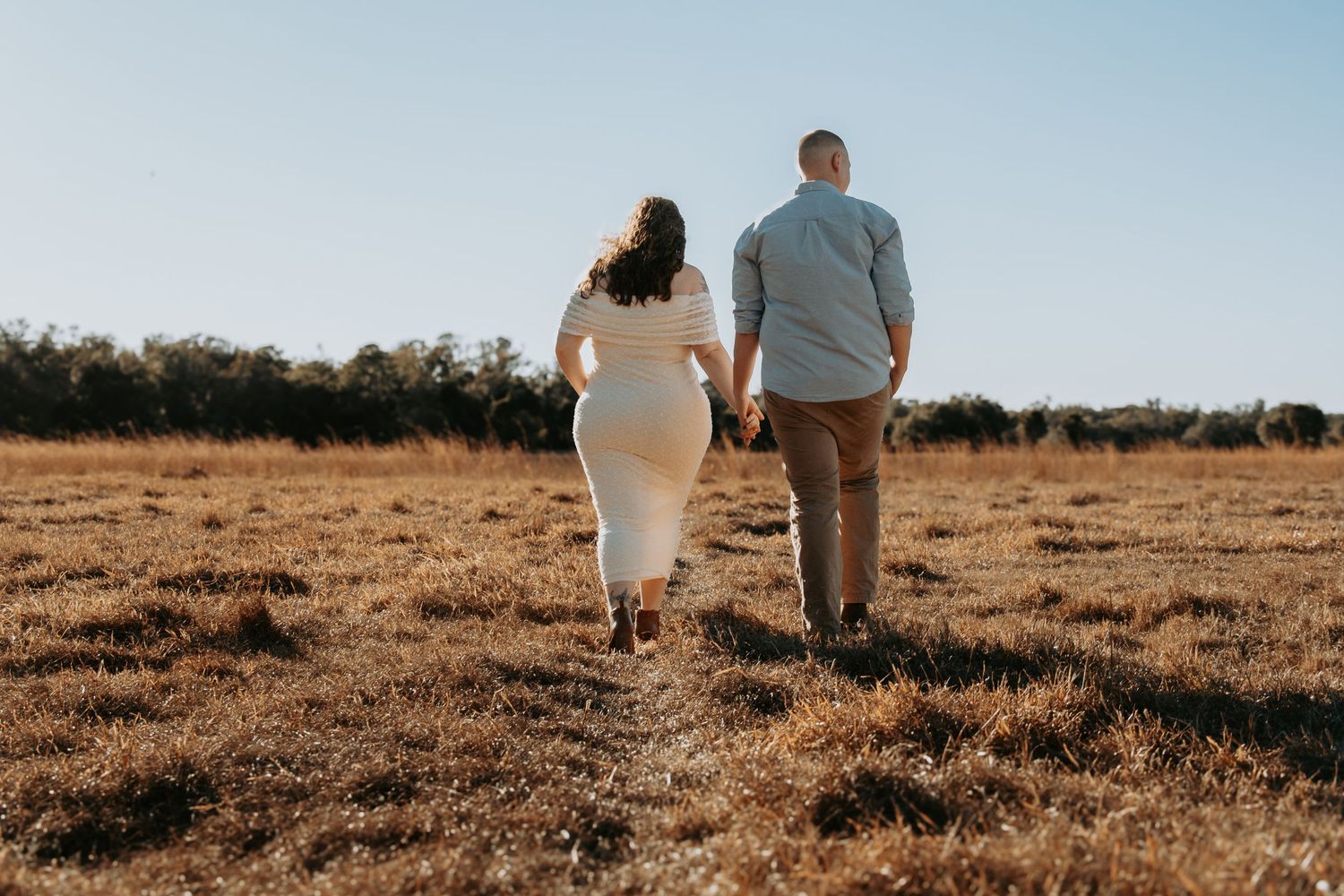 Couple holding hands walking through an open field with trees in the background on a sunny day.