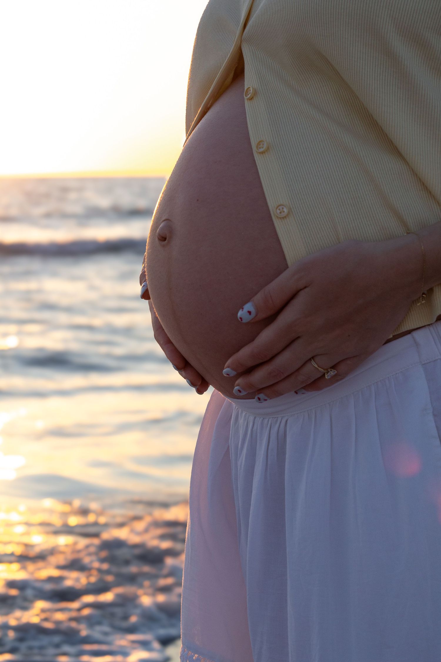 Close-up of pregnant woman cradling bare baby bump at golden sunset beach in white skirt.