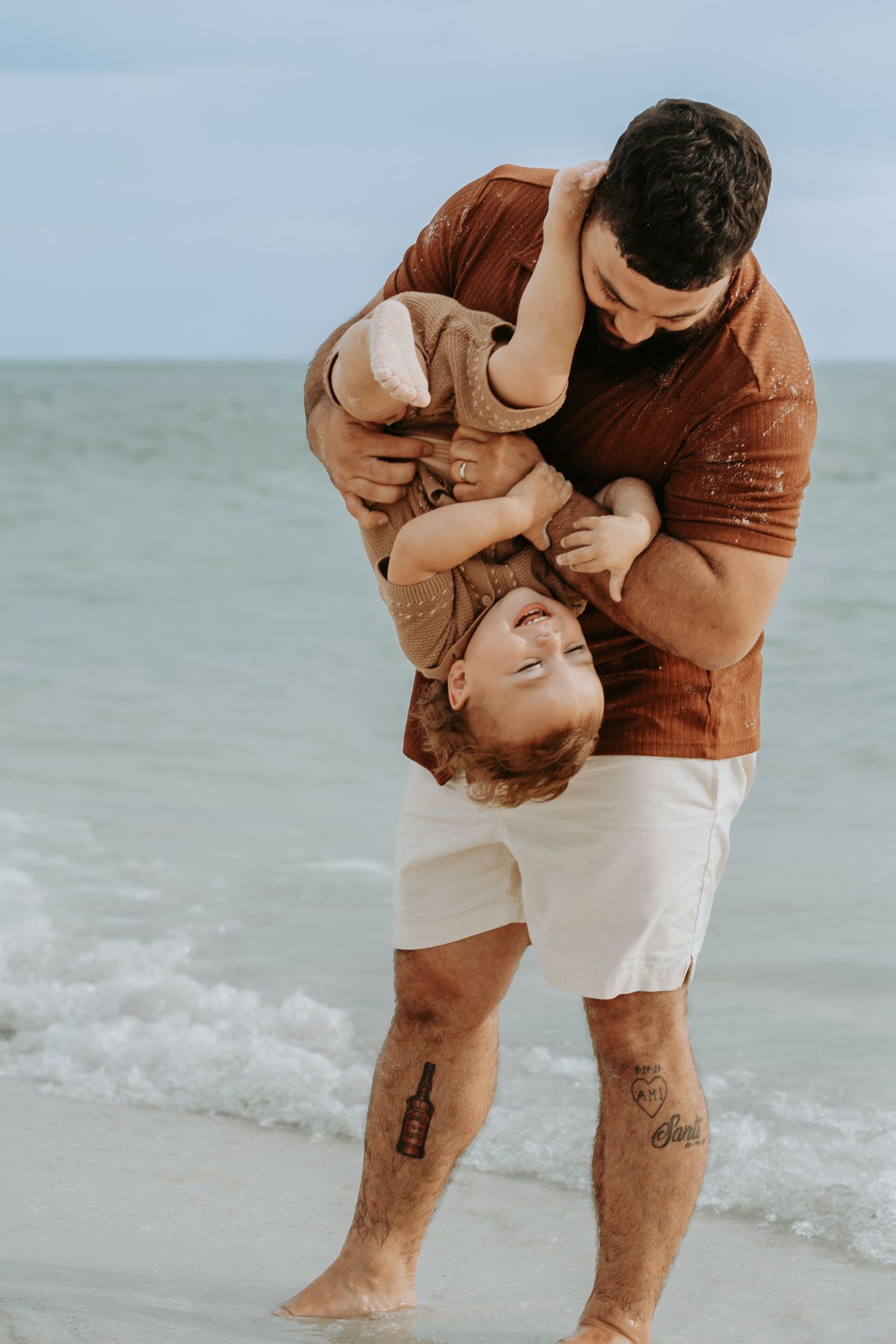 Father playfully holds laughing child upside down on a sandy beach with ocean waves in the background.