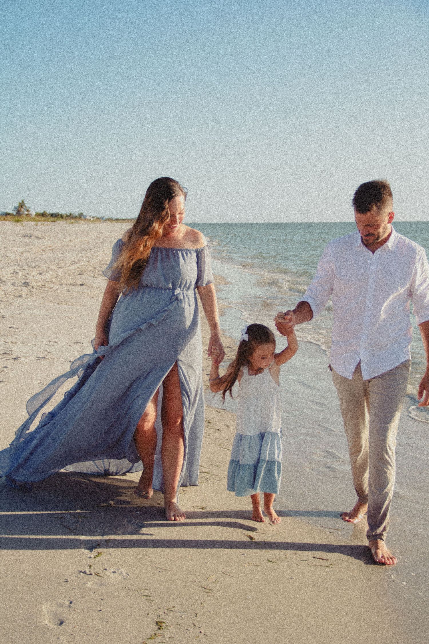 Family of three walking barefoot on sandy beach, parents in blue dress and white shirt swinging young daughter between them.