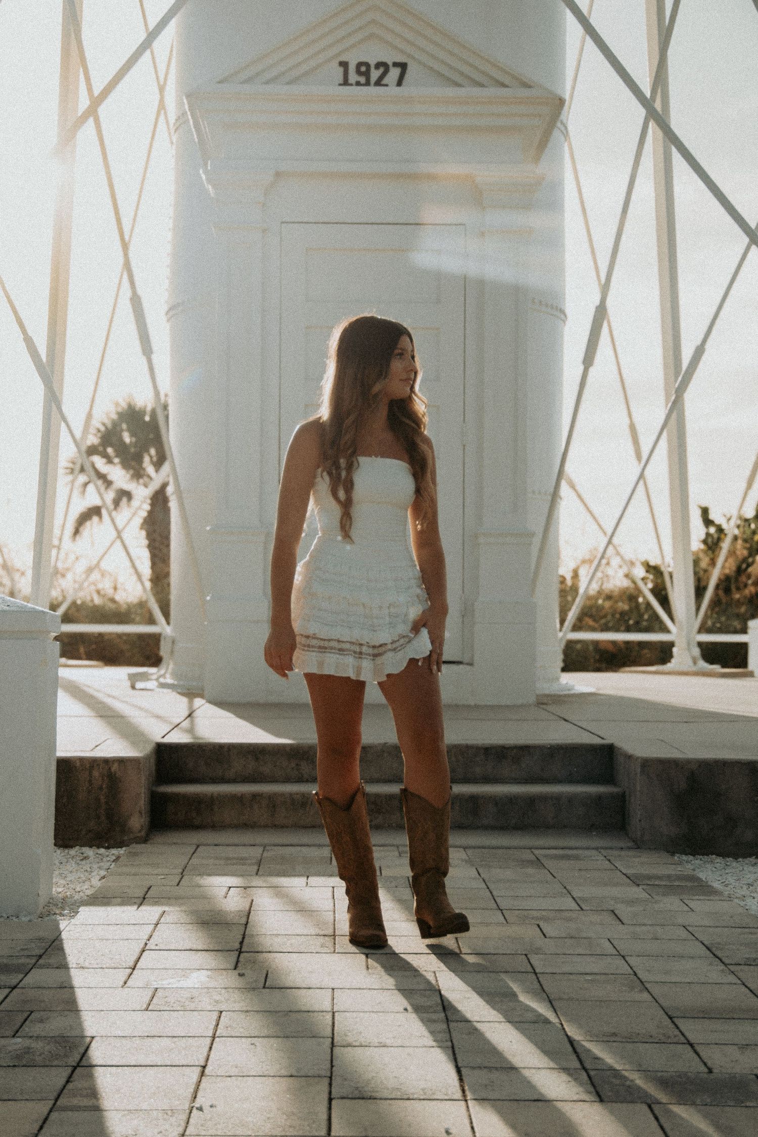 Young woman in white strapless mini dress and brown cowboy boots posing by a 1927 lighthouse.
