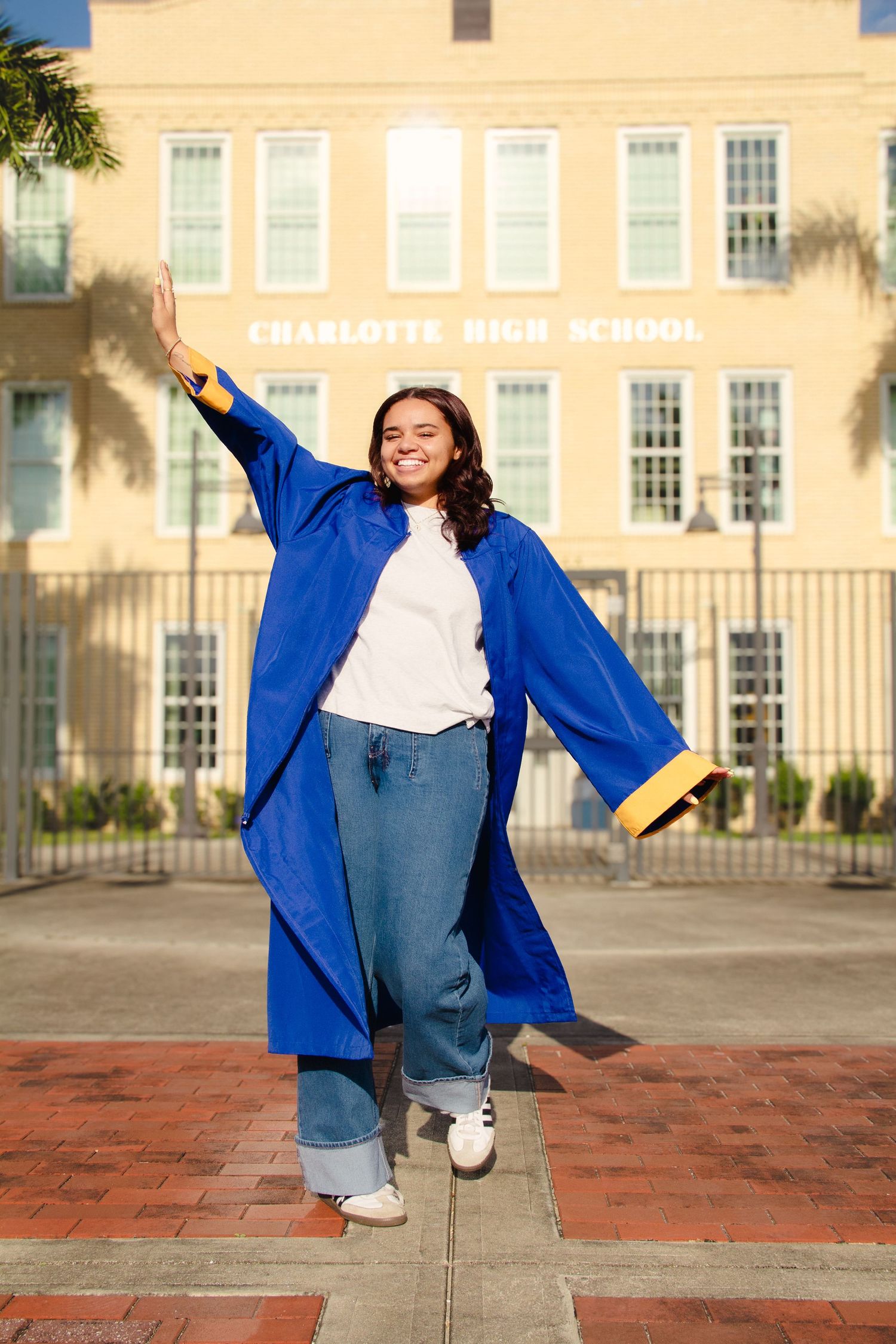 Smiling graduate in blue cap and gown poses in front of Charlotte High School building on a sunny day.
