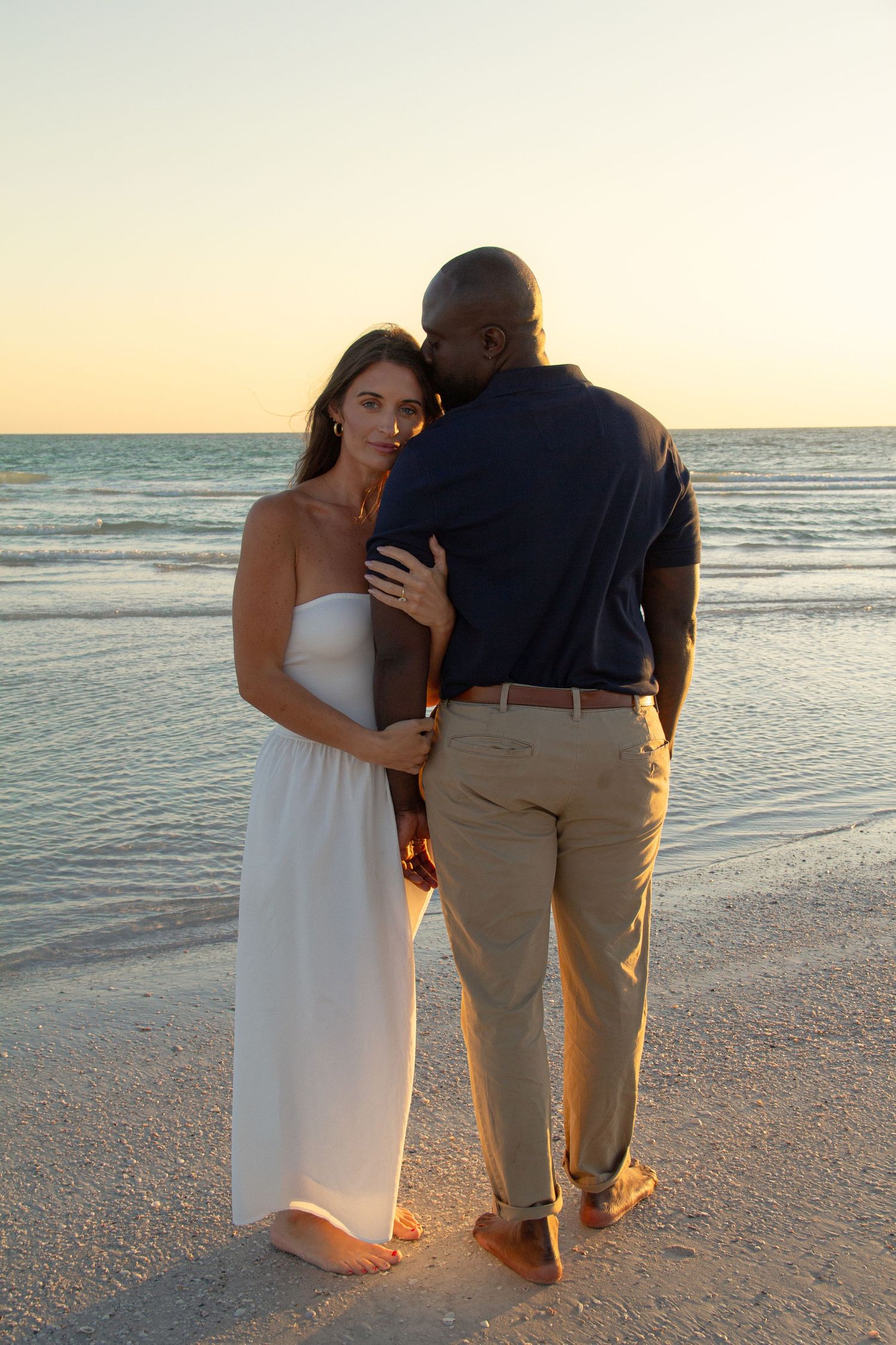 Interracial couple sharing a romantic moment on a sandy beach at sunset, woman in white dress, man in navy shirt.