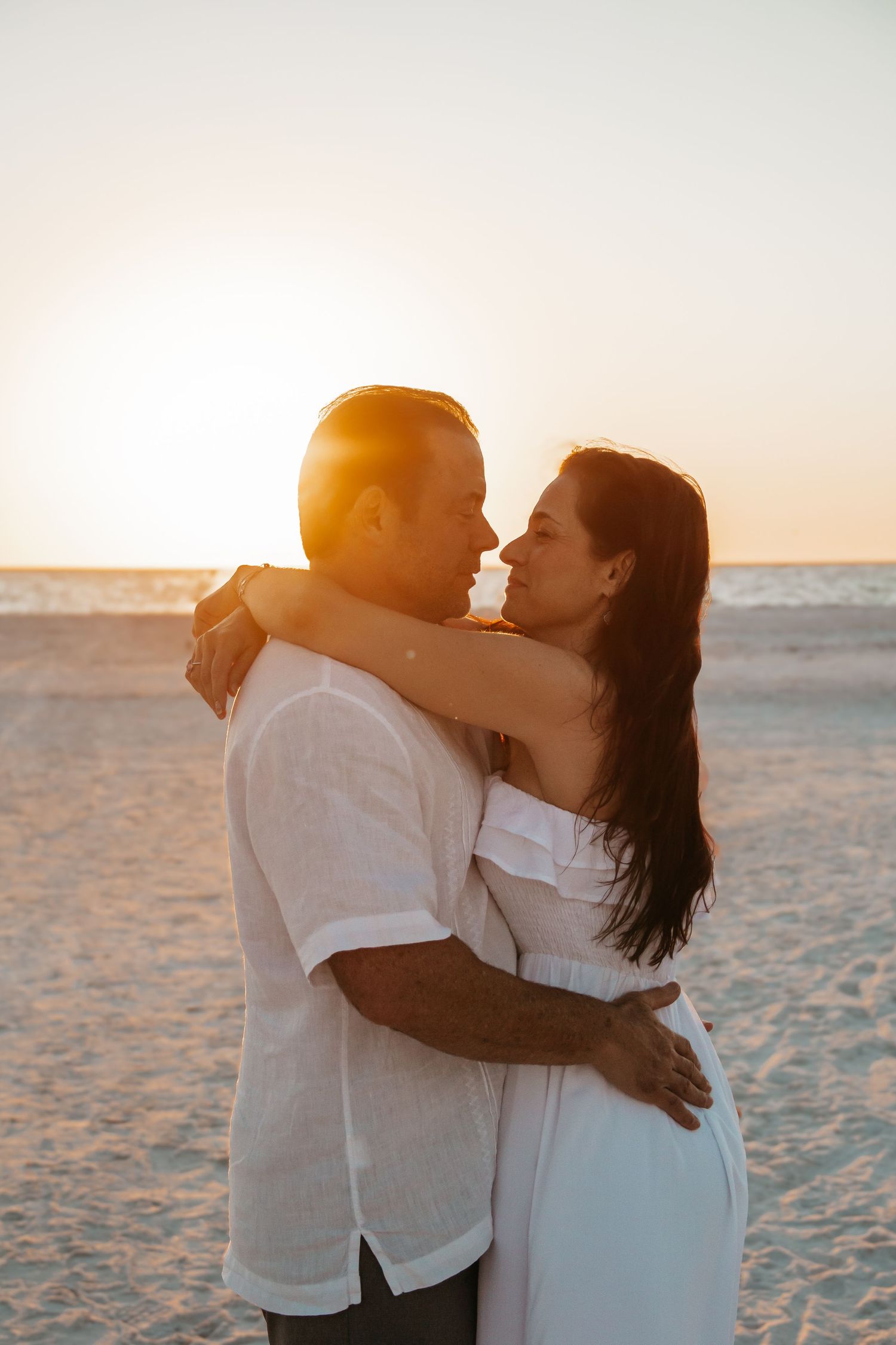 Couple embracing romantically on a sandy beach at golden sunset, wearing white outfits, sharing a tender moment.