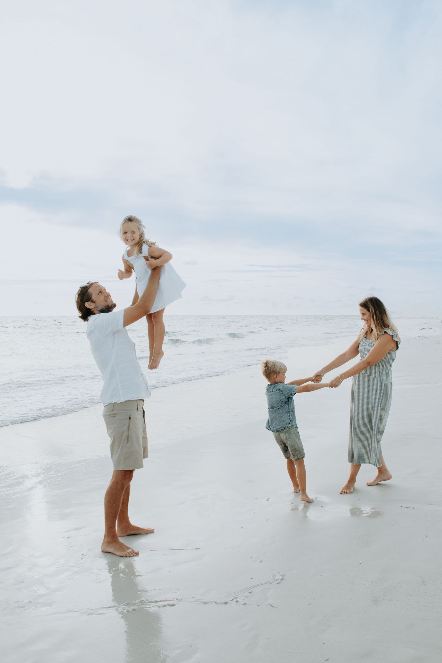 Family of four playing on a beach, father lifting daughter while mother holds son's hands near ocean waves.