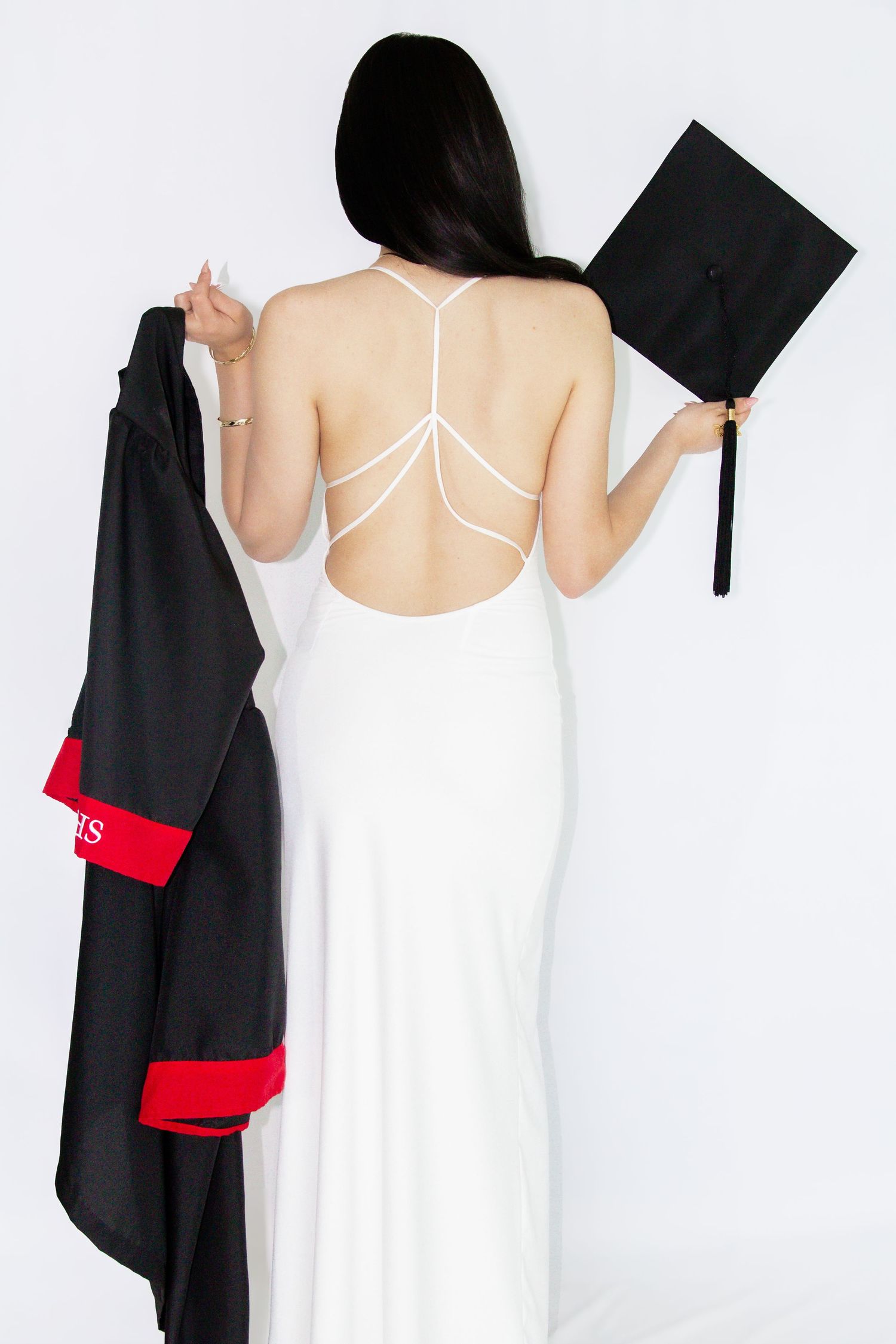 Woman in backless white gown holding graduation cap and black robe against white background.