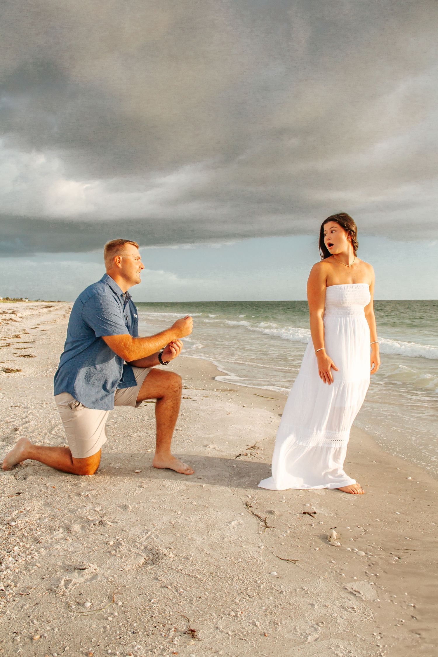Man proposes on one knee to surprised woman in white dress on a sandy beach with dramatic cloudy sky.