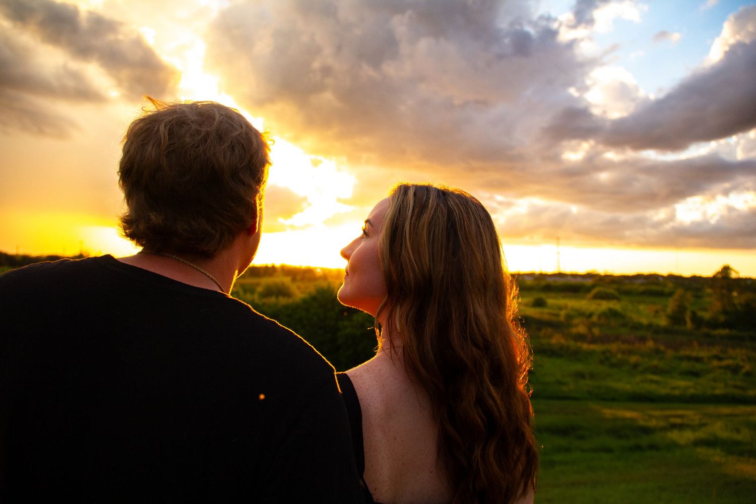 Couple watching a dramatic golden sunset over lush green landscape with cloudy sky.