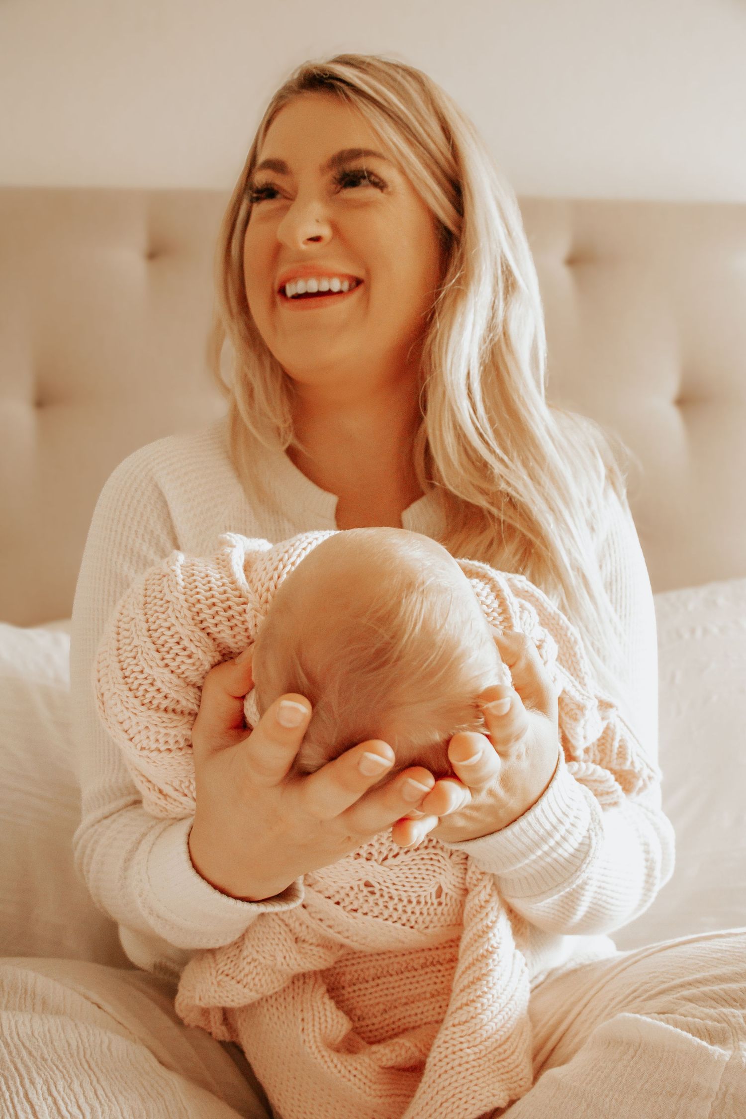 Smiling blonde woman in white sweater holding newborn baby in pink knit blanket while sitting on bed.