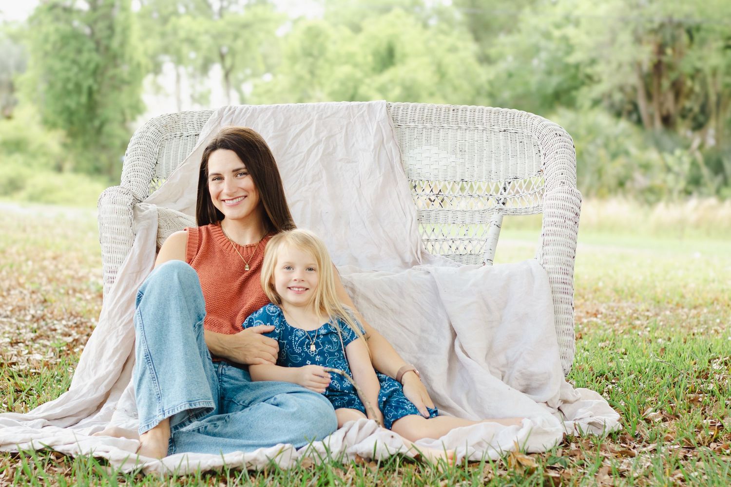 Woman and young blonde girl smiling together on a white wicker bench outdoors on green grass.