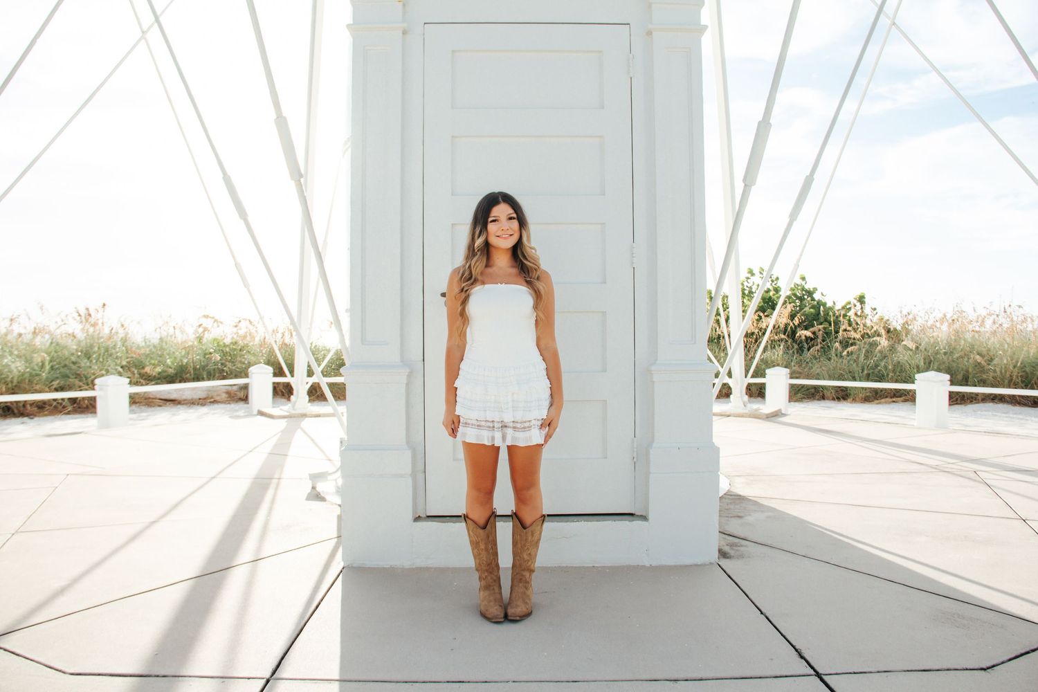 Woman in white romper and brown boots standing before a white door on a bright sunny day.
