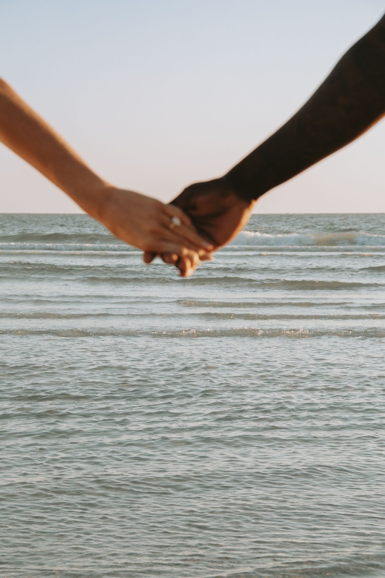 Close-up of interracial couple holding hands at the beach with ocean waves in the background.