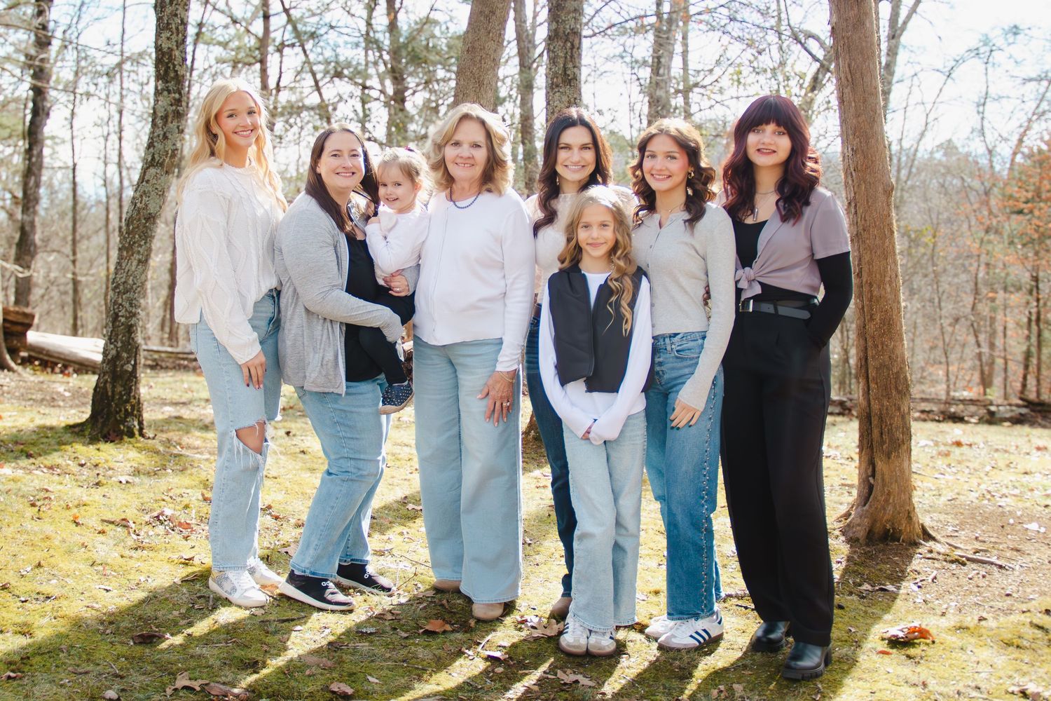 Seven women and girls pose together outdoors in a wooded area, wearing casual outfits in neutral tones.