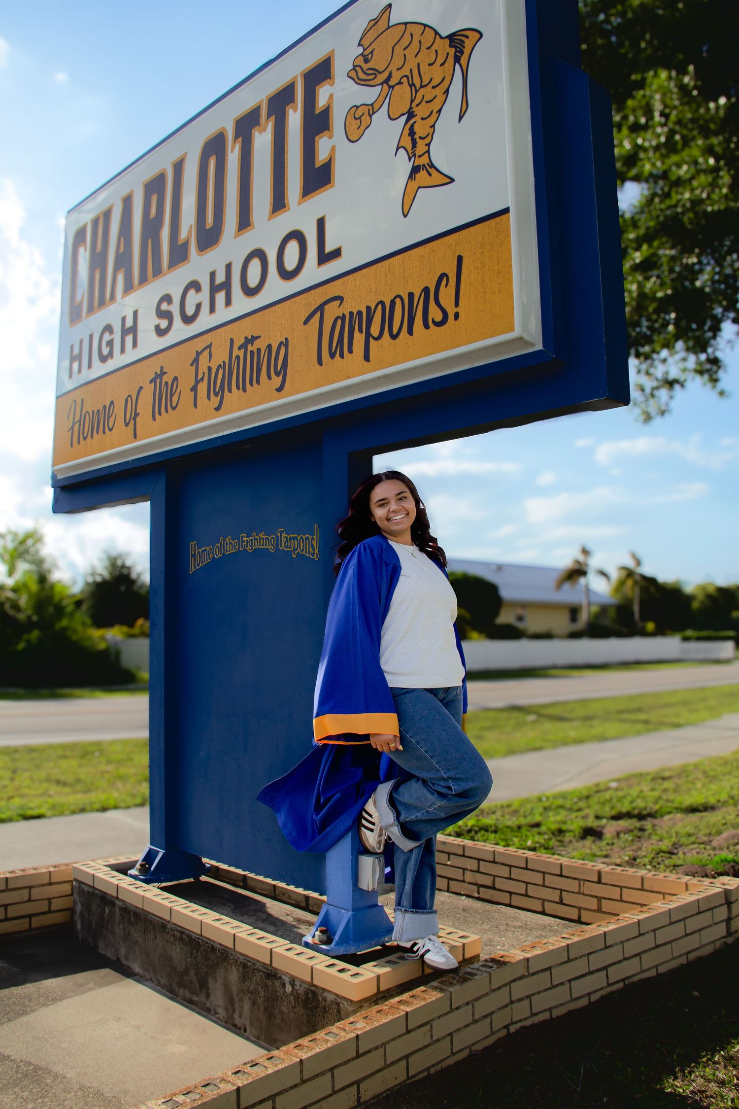 A smiling senior student poses in graduation cap and gown beside Charlotte High School's Fighting Tarpons sign.