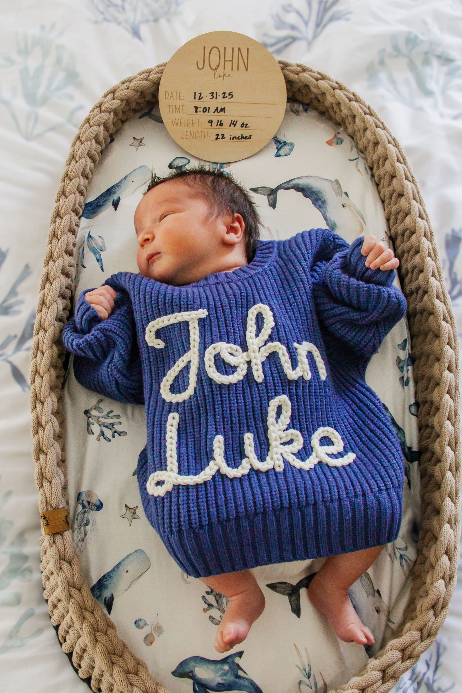 Newborn baby sleeping in a wicker Moses basket wearing a navy knit sweater embroidered with John Luke.