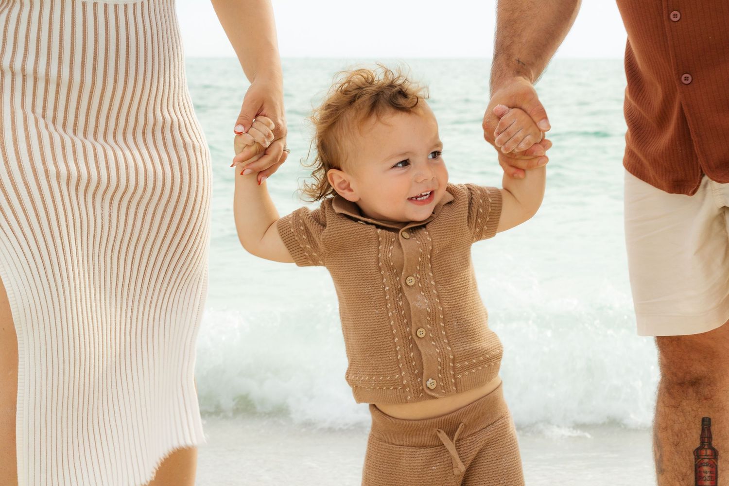 A happy toddler in a beige knit outfit holds parents hands while learning to walk on a sunny beach.