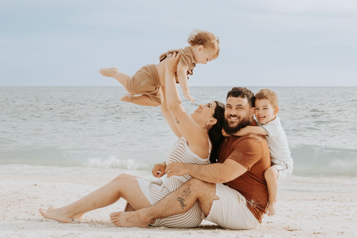 Family of four enjoying a beach day, parents lifting laughing toddlers on sandy shore with ocean backdrop.