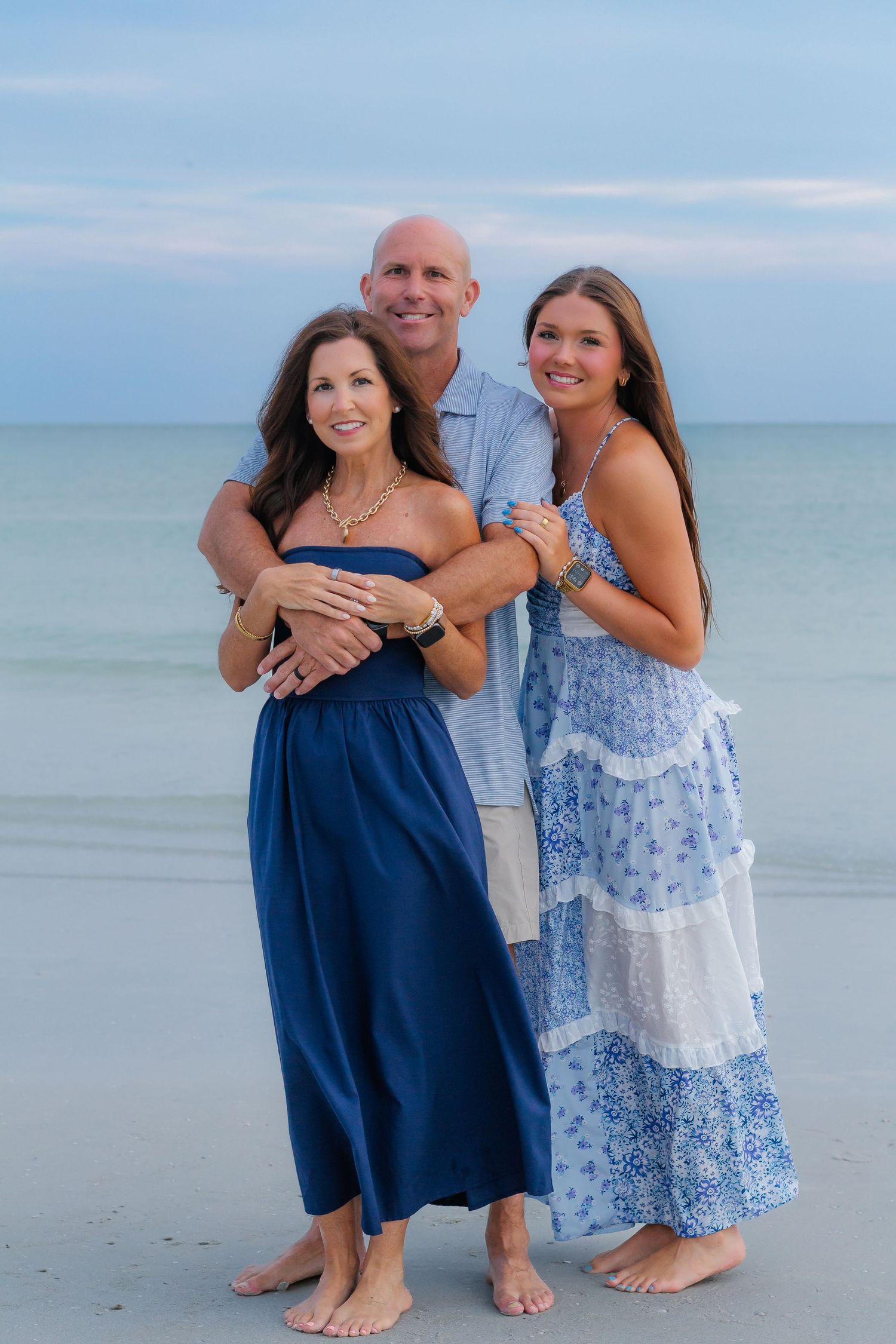 Family of three poses barefoot on a sandy beach in coordinating blue outfits with calm ocean waters in the background.